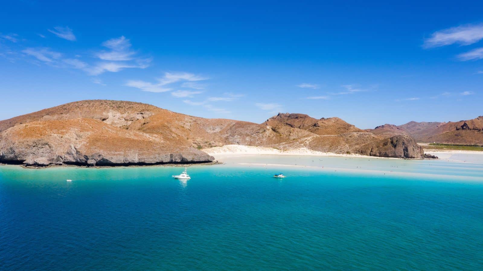 Aerial view of a beach with turquoise water, white sand, and rocky brown hills in the background. Three boats are floating near the shore under a clear blue sky.