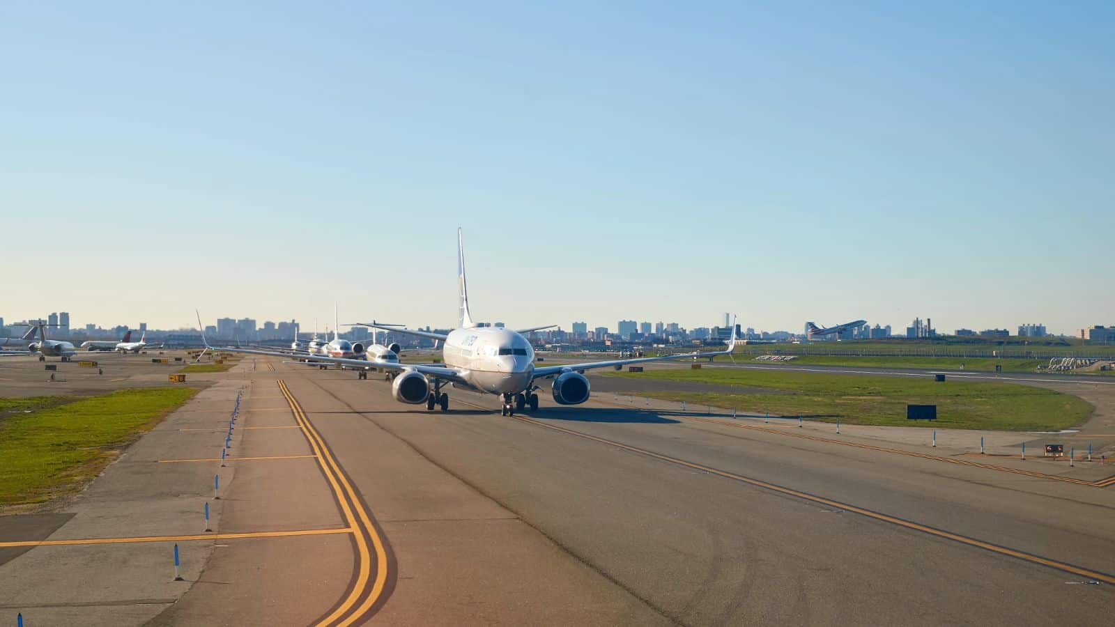An airplane taxis on an airport runway with several other planes waiting in line behind it. The sky is clear, and a city skyline is visible in the background.