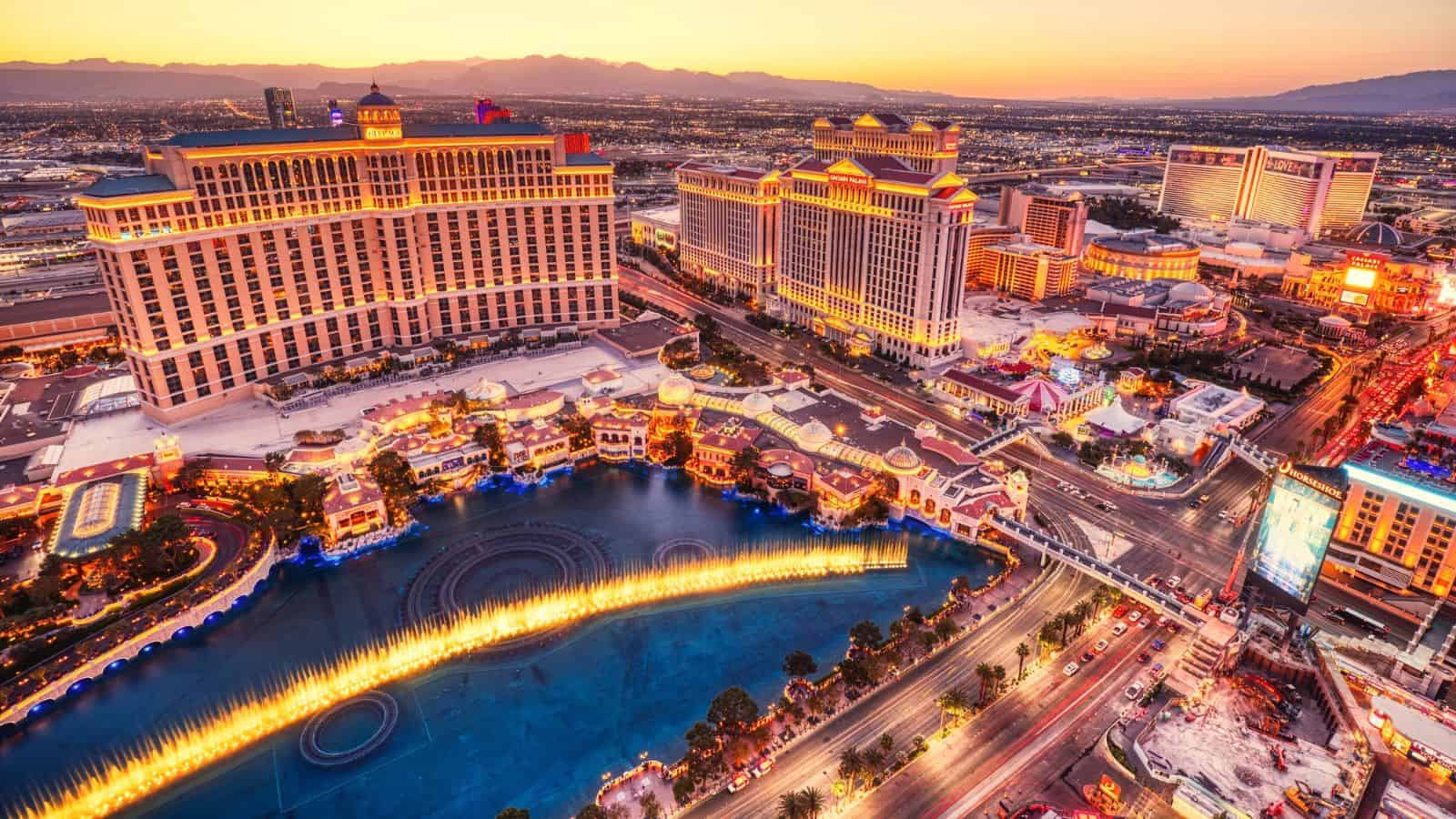 Aerial view of the Las Vegas Strip at sunset showing hotels, casinos, and the Bellagio fountains performing, with city lights and distant mountains in the background.