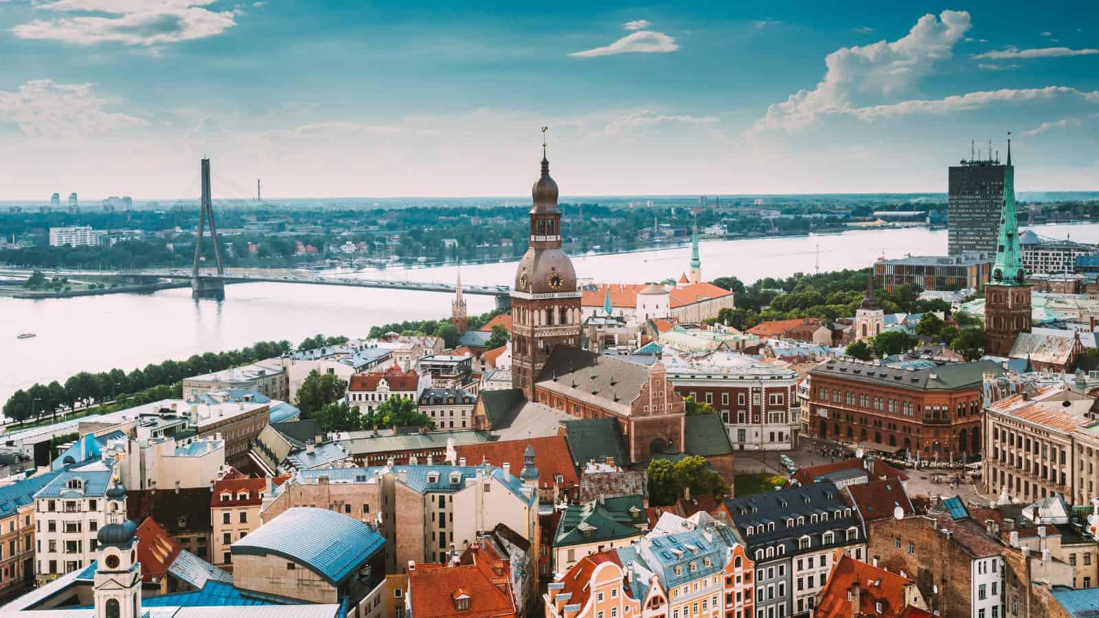 A panoramic view of Riga, Latvia, showing the Daugava River, the cathedral with a tall spire in the center, red-roofed buildings, and a cable-stayed bridge in the background under a partly cloudy sky.