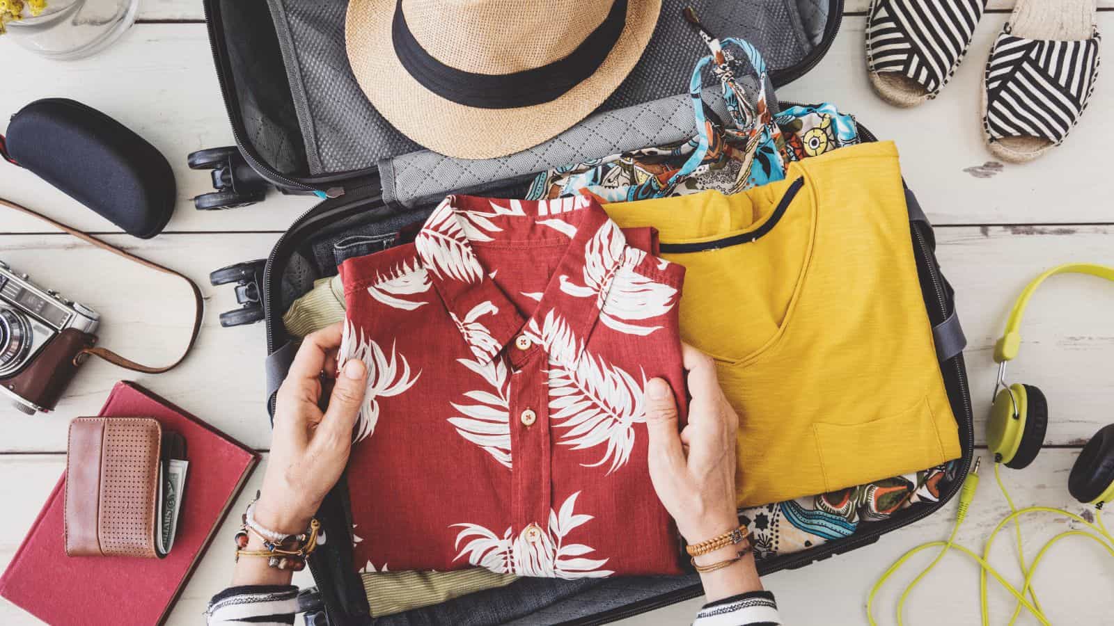 A person packs a suitcase with a red floral shirt, yellow top, and other clothes. Surrounding items include a hat, sunglasses, striped shoes, camera, book, wallet, and headphones on a wooden surface.