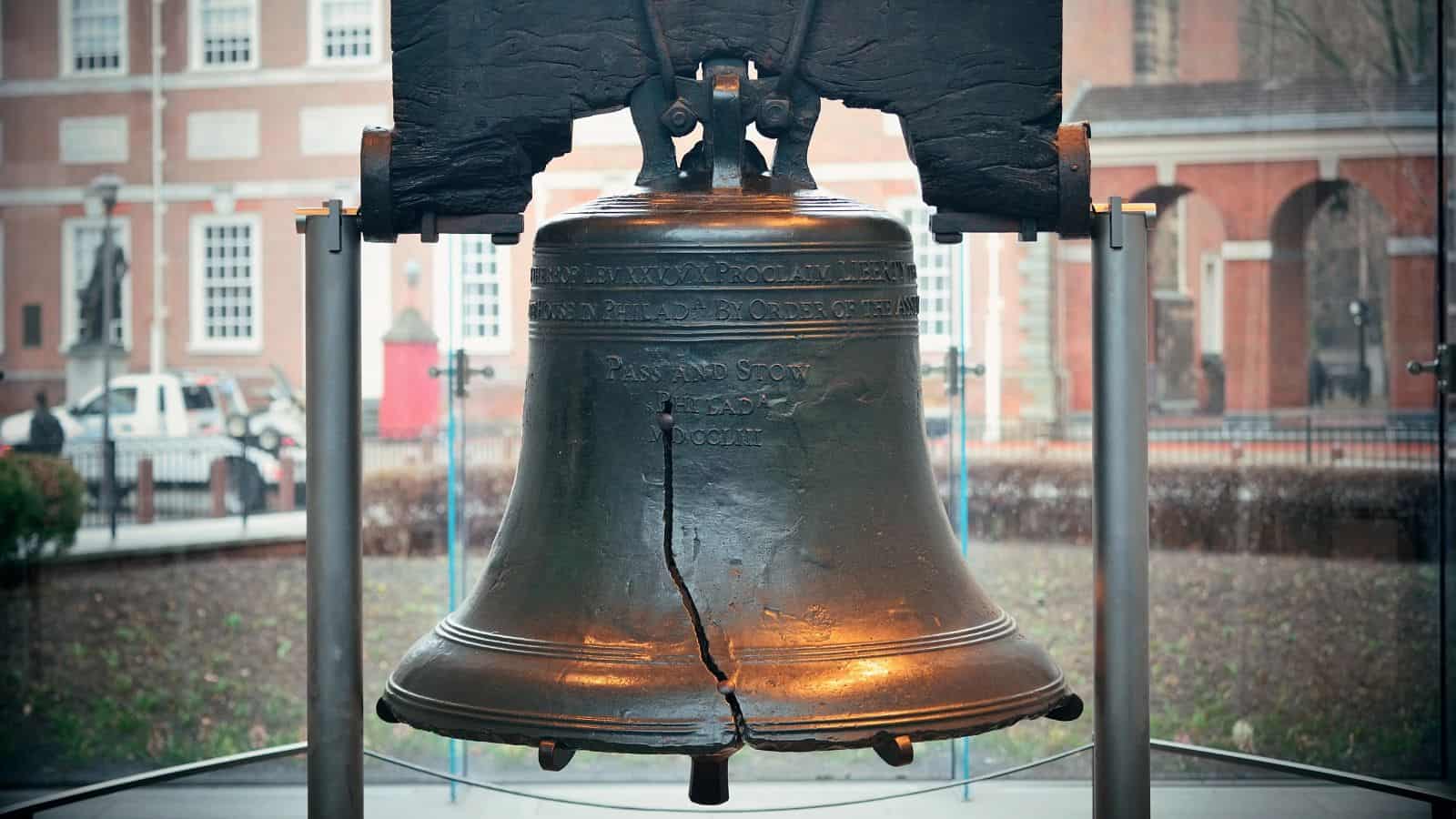 A large, cracked bronze bell known as the Liberty Bell is displayed indoors behind protective barriers, with a brick building and windows visible in the background.