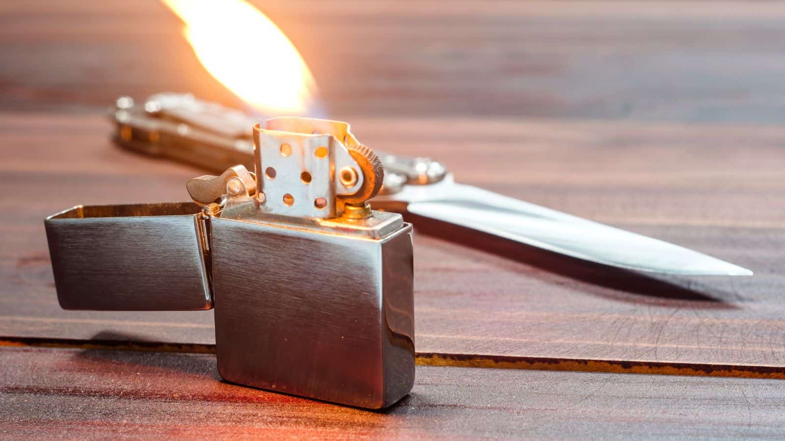 A metal flip-top lighter with an open lid and flame is on a wooden surface, with a folding knife lying in the background.