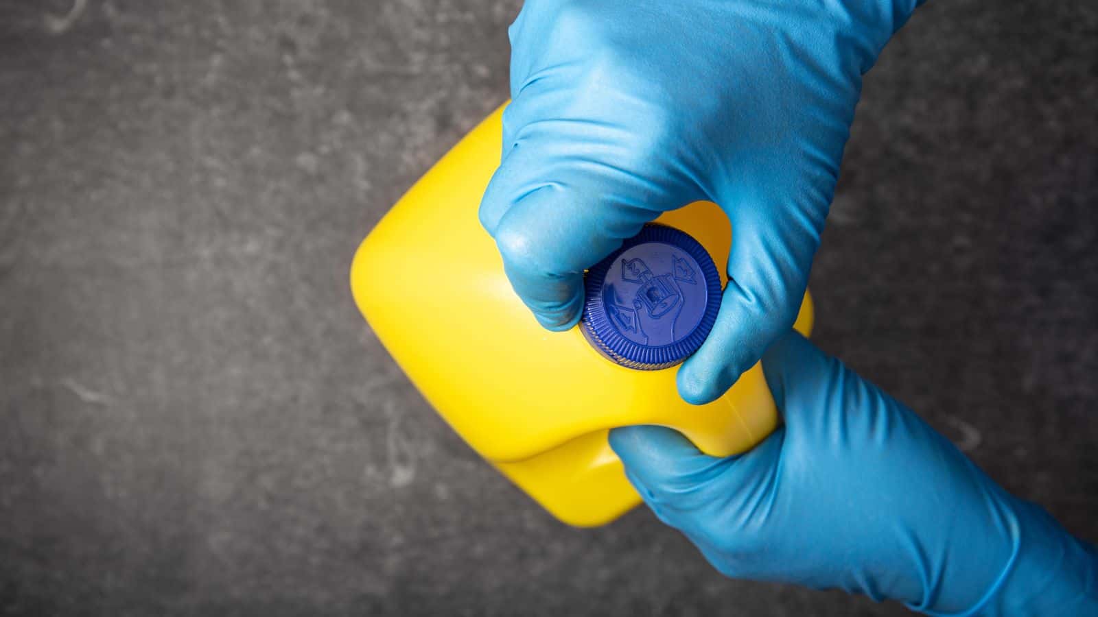 Gloved hands are opening the blue cap of a yellow plastic container, likely containing cleaning chemicals, against a dark, textured background.
