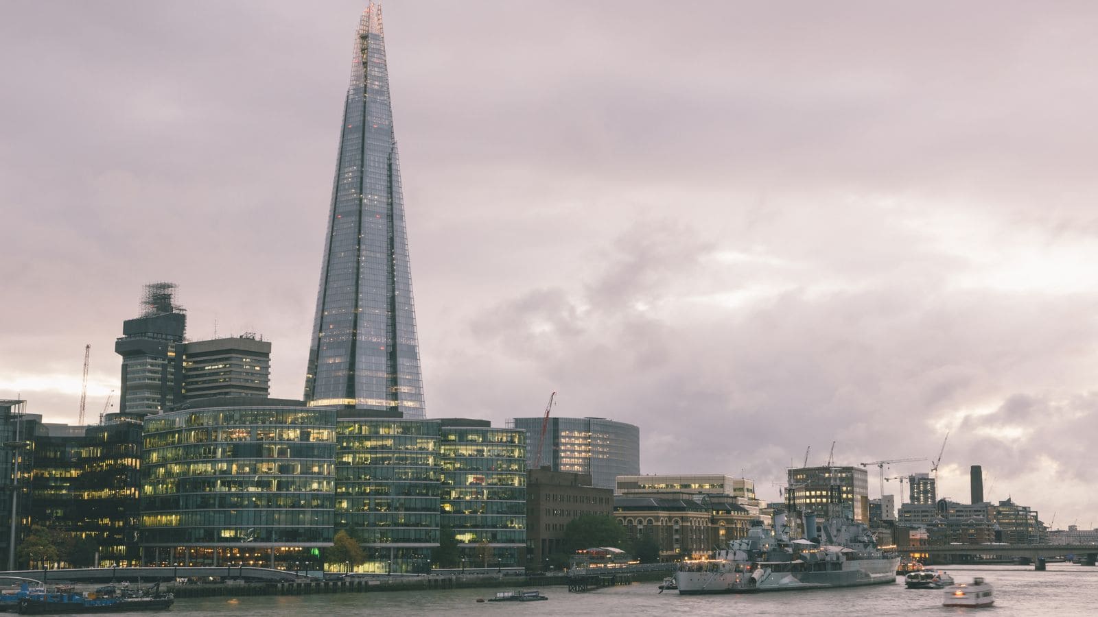 A view of The Shard skyscraper in London at dusk, with office buildings lit up, the River Thames in the foreground, and boats on the water under a cloudy sky.