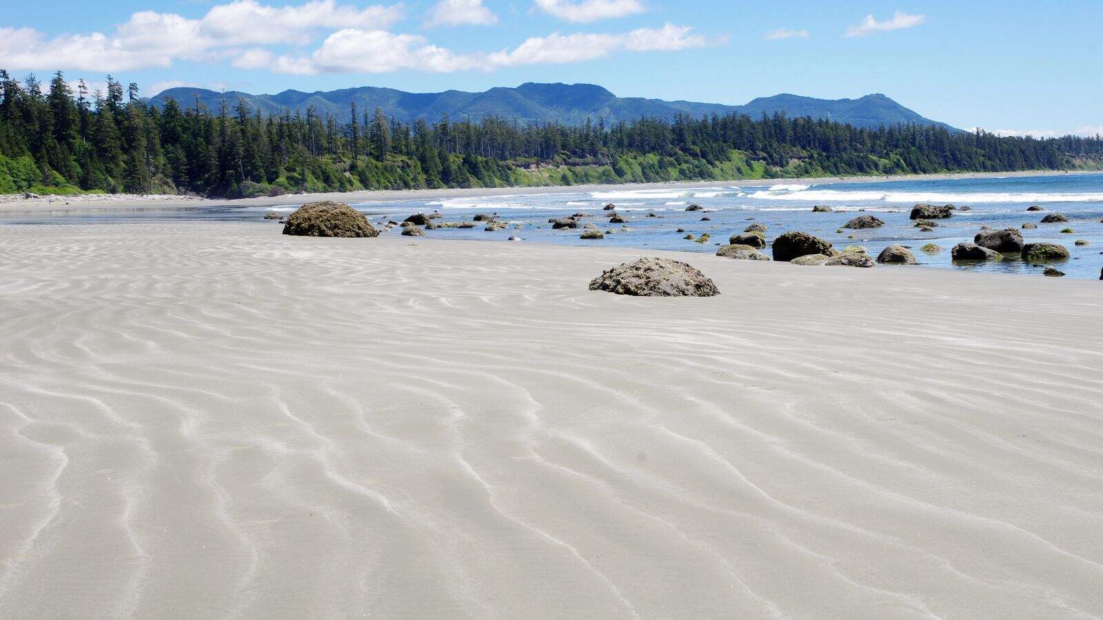 Wide sandy beach with rippled patterns in the sand, scattered rocks, gentle waves, tree-lined shore, and forested mountains in the background under a partly cloudy sky.