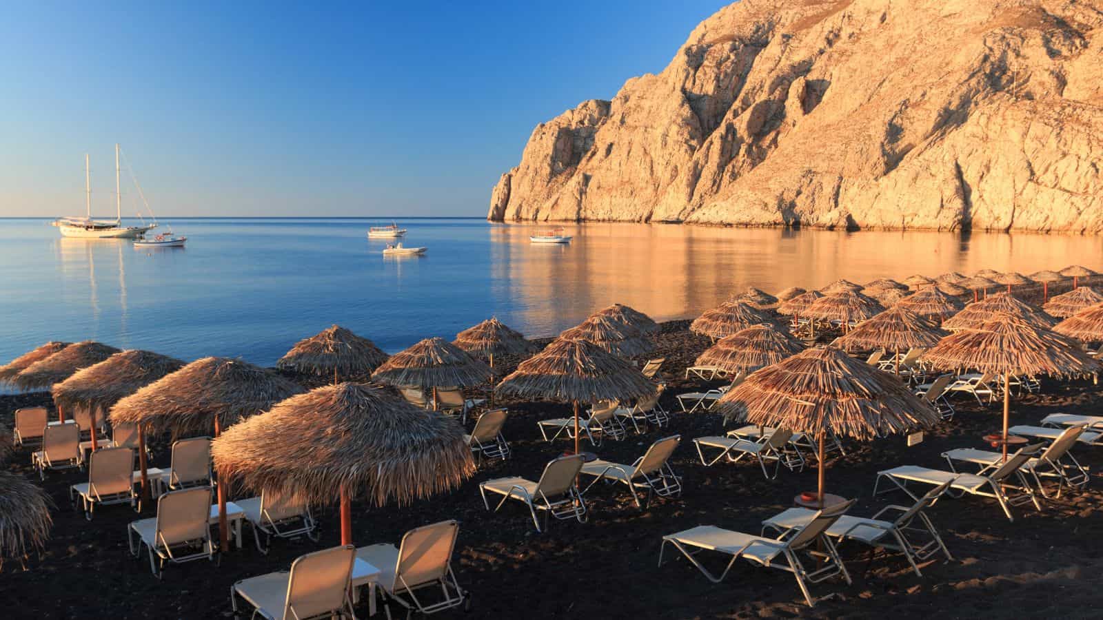 Rows of empty lounge chairs and straw umbrellas on a black sand beach face calm blue water with anchored boats; a rocky cliff is visible in the background under a clear sky.