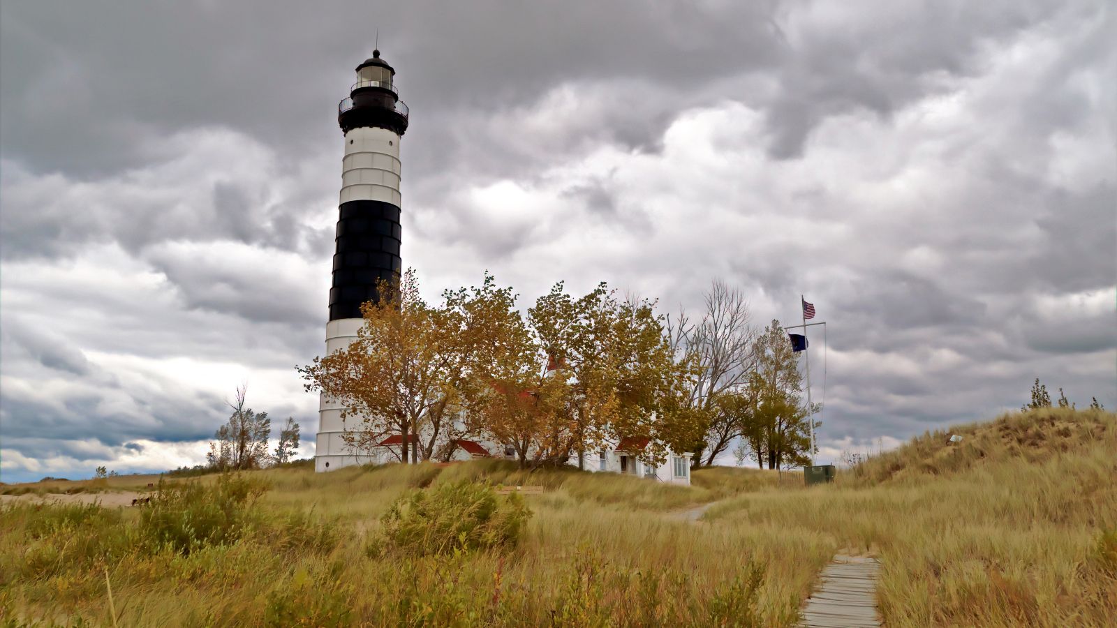 A black and white striped lighthouse stands behind autumn trees and tall grass under a cloudy sky, with a flagpole nearby and a wooden path leading toward the structure.