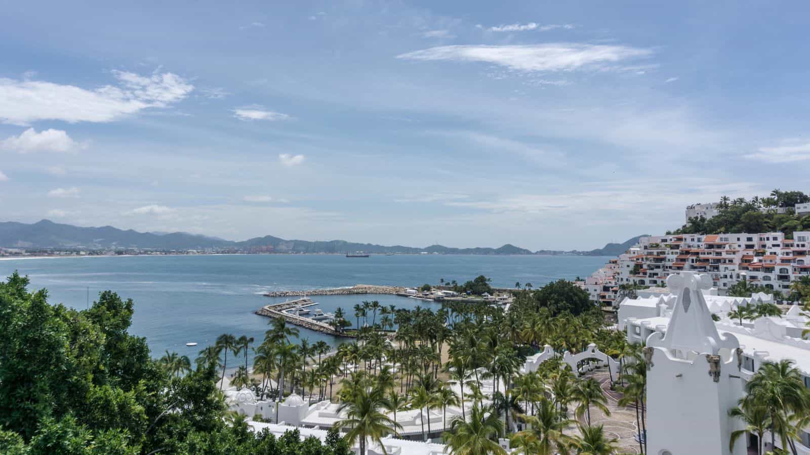 A coastal scene featuring a marina with a breakwater, palm trees, and white buildings on a hillside overlooking a calm bay with mountains in the background under a partly cloudy sky.