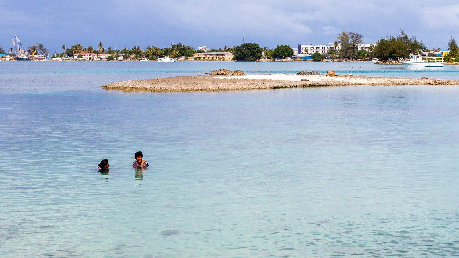 Two people swim in calm, shallow water near a sandy island—a serene scene that echoes the beauty of vanishing countries threatened by rising seas. In the background are buildings, trees, and boats along the shoreline under a cloudy sky.