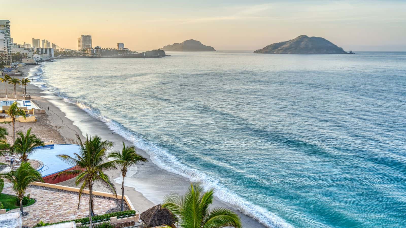 View of a sandy beach lined with palm trees, a swimming pool near the shore, calm ocean waves, distant city buildings on the left, and two large islands visible on the horizon under a clear sky at sunset.