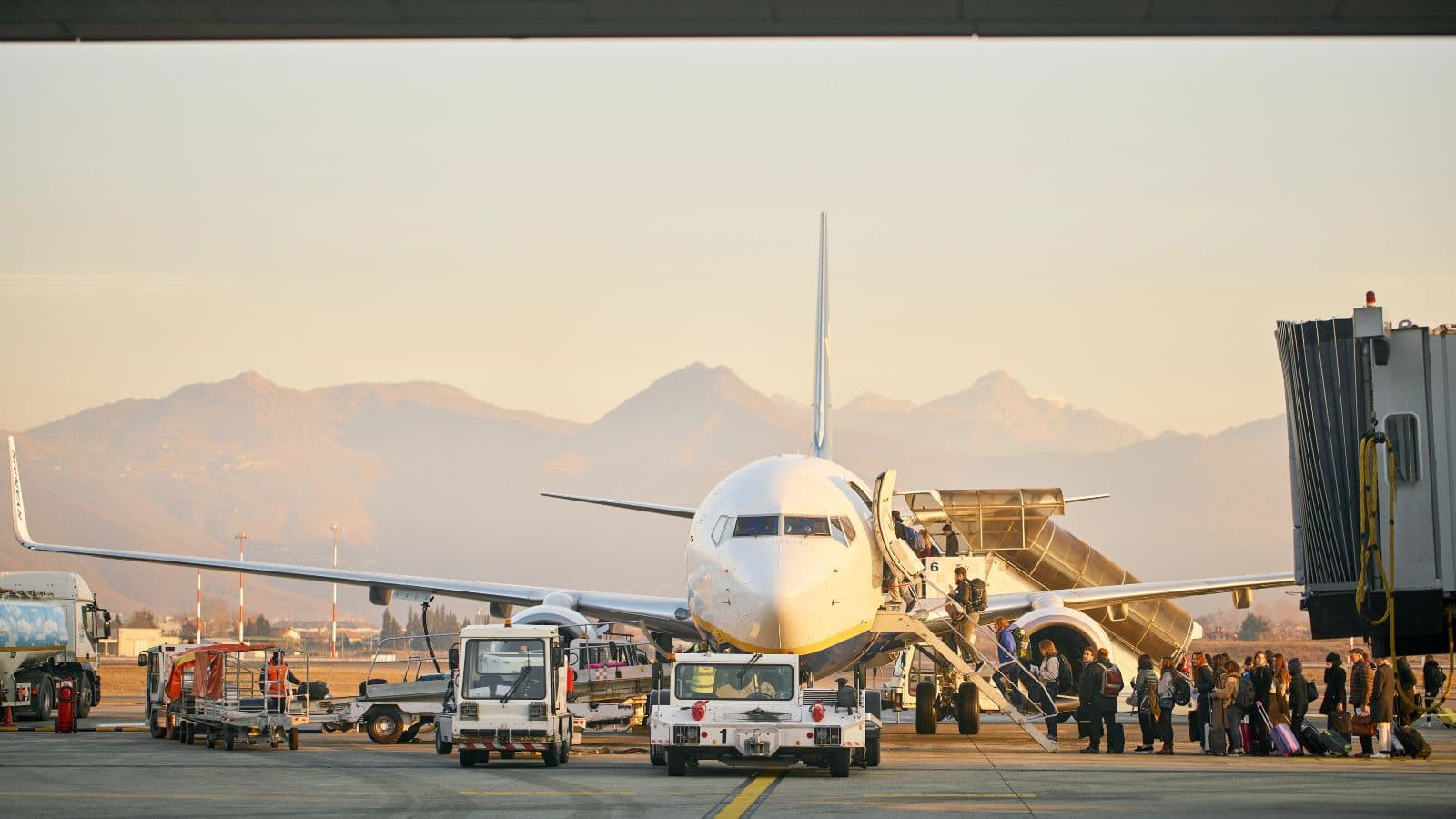 A commercial airplane is parked at an airport gate with boarding stairs attached. Passengers are boarding and airport vehicles and ground crew are near the plane. Mountains are visible in the background under a clear sky.