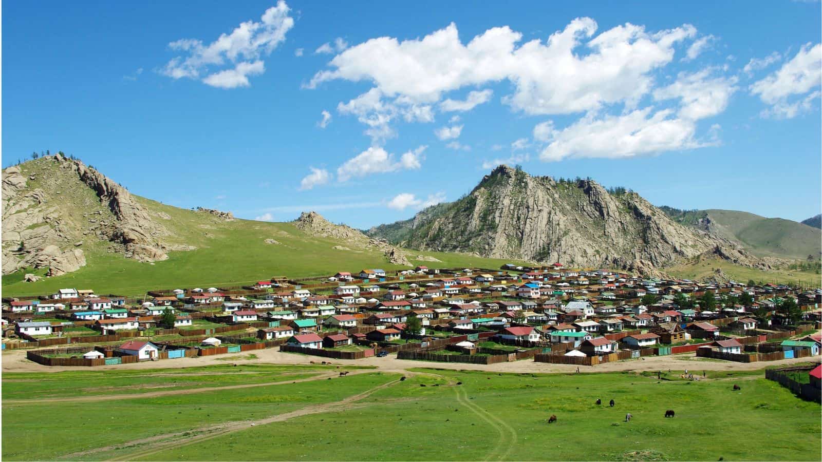 A rural village with many small houses and colorful roofs is spread across a grassy hillside, with rocky mountains in the background under a blue sky with scattered clouds.