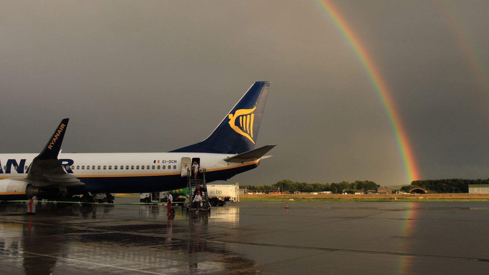 A Ryanair airplane is parked on a wet airport tarmac under a dark sky with a visible rainbow in the background. Passengers are boarding the plane using stairs at the rear. Trees and buildings are in the distance.