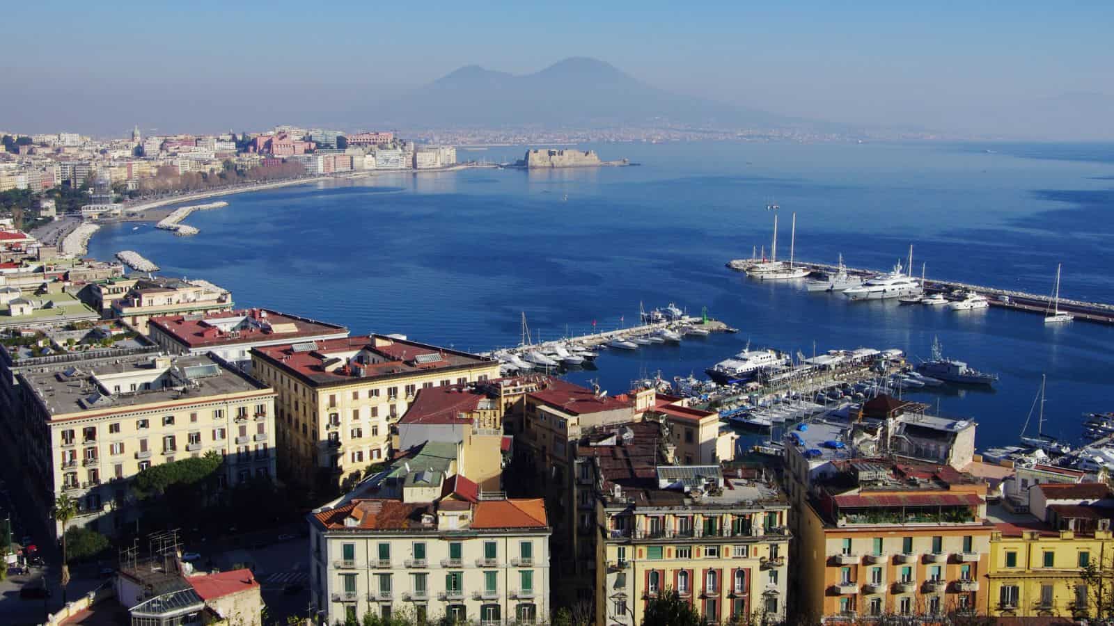 A coastal city with colorful buildings, a marina with yachts, and a curved bay. Mount Vesuvius is visible in the background under a clear blue sky.