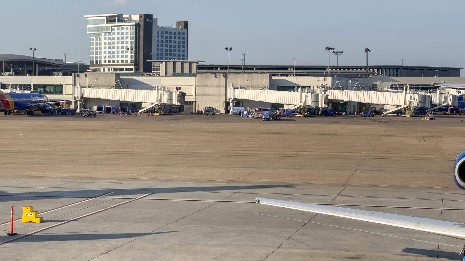 A view of an airport terminal with multiple jet bridges connected to airplanes. Ground vehicles and equipment are nearby, and a tall modern building is visible in the background under a clear sky.