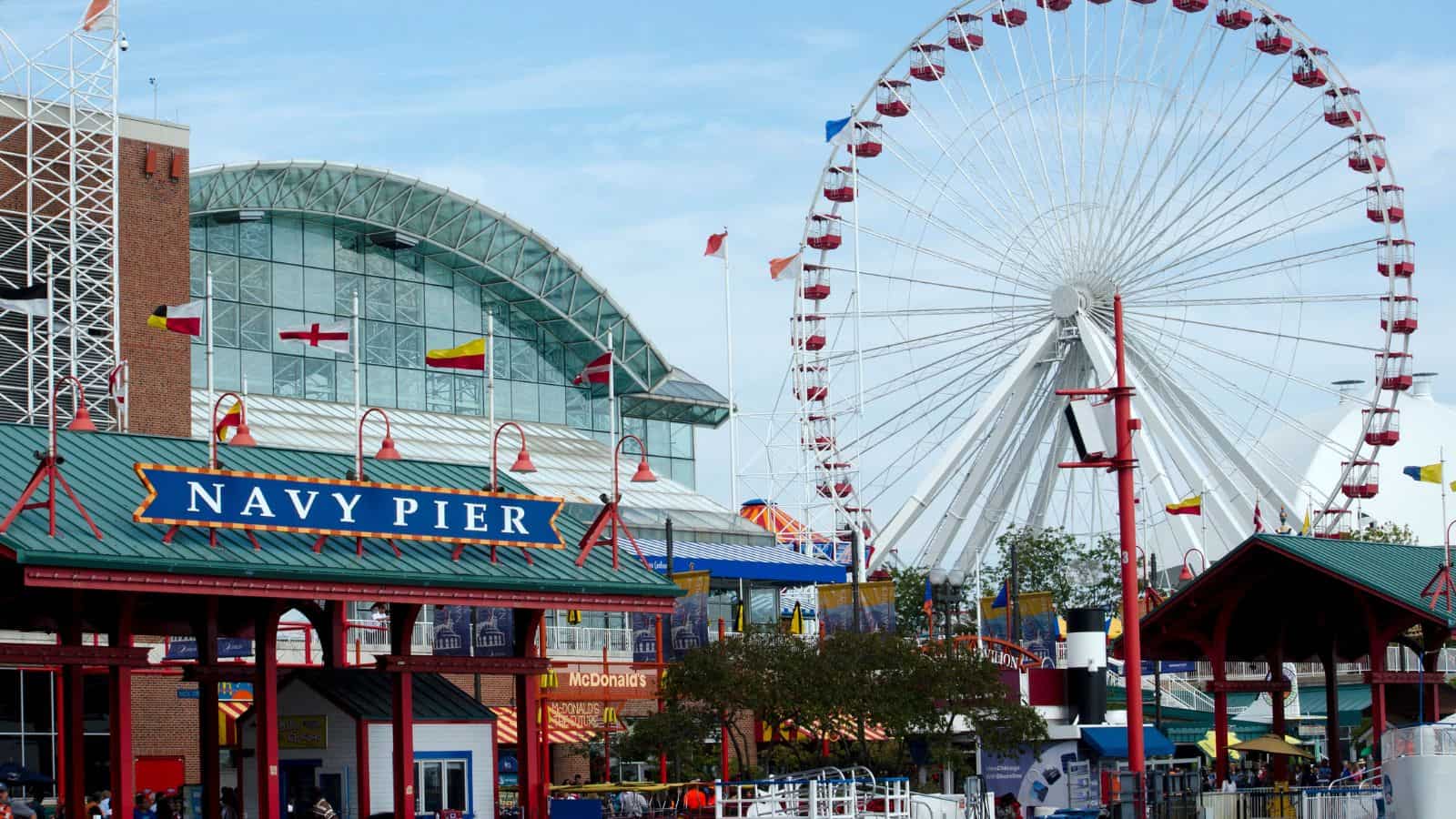 Navy Pier entrance with a large Ferris wheel, colorful flags, and a glass building in the background. Several people are visible near the entrance and a McDonald's sign appears below the glass building.