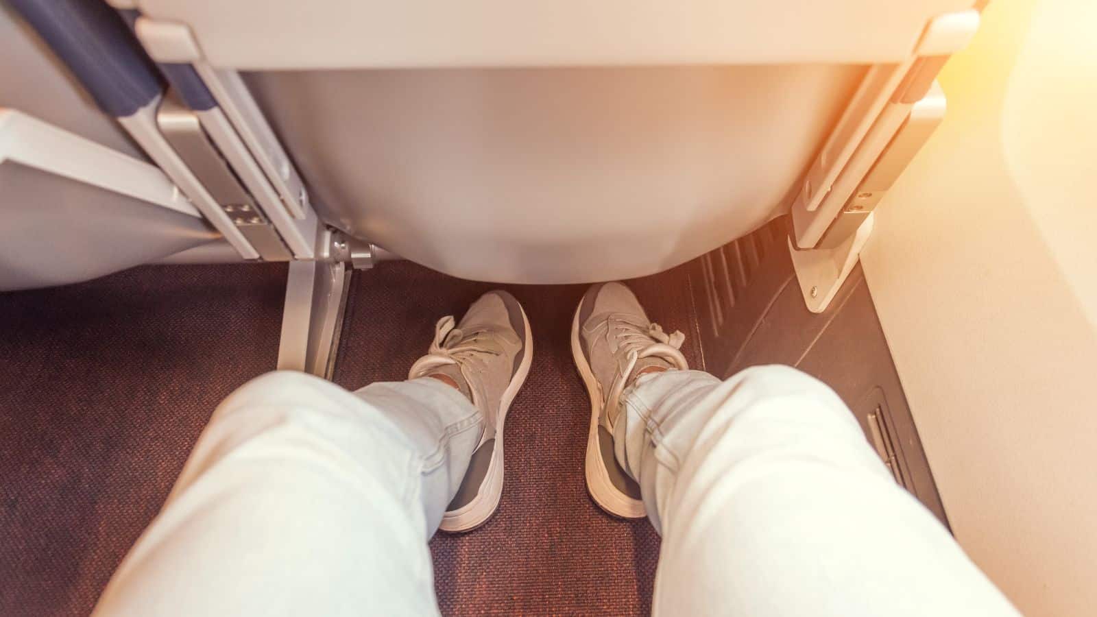 A person seated on an airplane with legs extended, showing their sneakers and light-colored pants—capturing a typical flight experience from their perspective, facing the seat in front and the airplane floor.