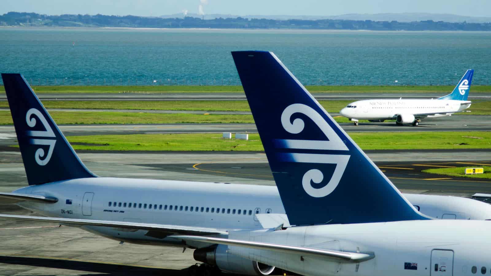 Two Air New Zealand planes are parked at an airport with a third Air New Zealand plane taxiing on the runway in the background. The ocean and distant land are visible beyond the airport.