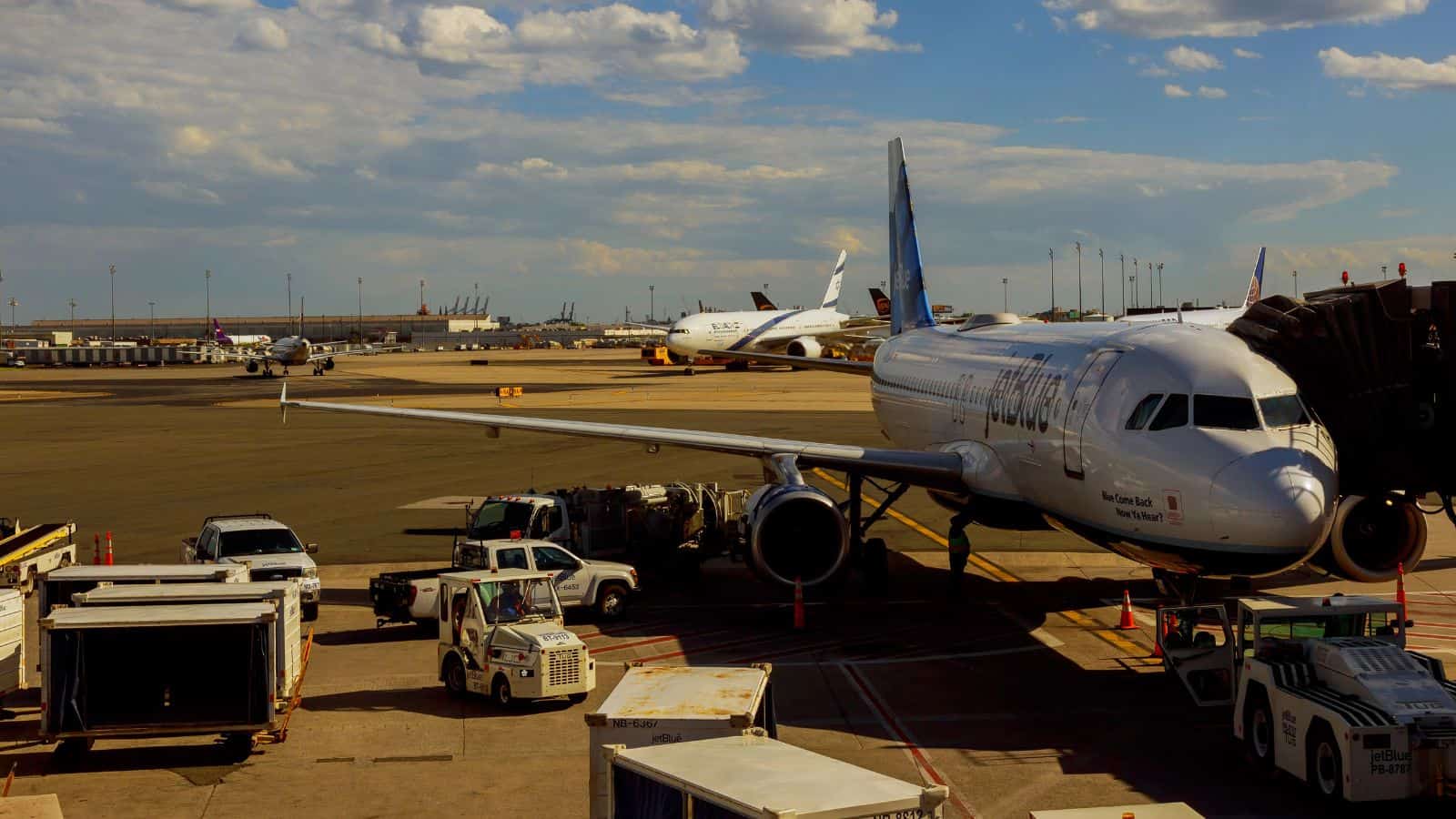A commercial airplane is parked at an airport gate with ground service vehicles around it. Other planes and airport buildings are visible in the background under a partly cloudy sky.