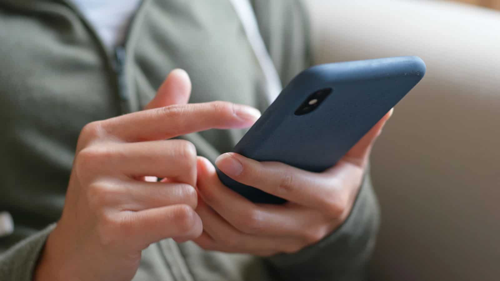 A person wearing a green zip-up jacket holds a smartphone with both hands, using their right index finger to tap the screen. The phone has a dark-colored case.