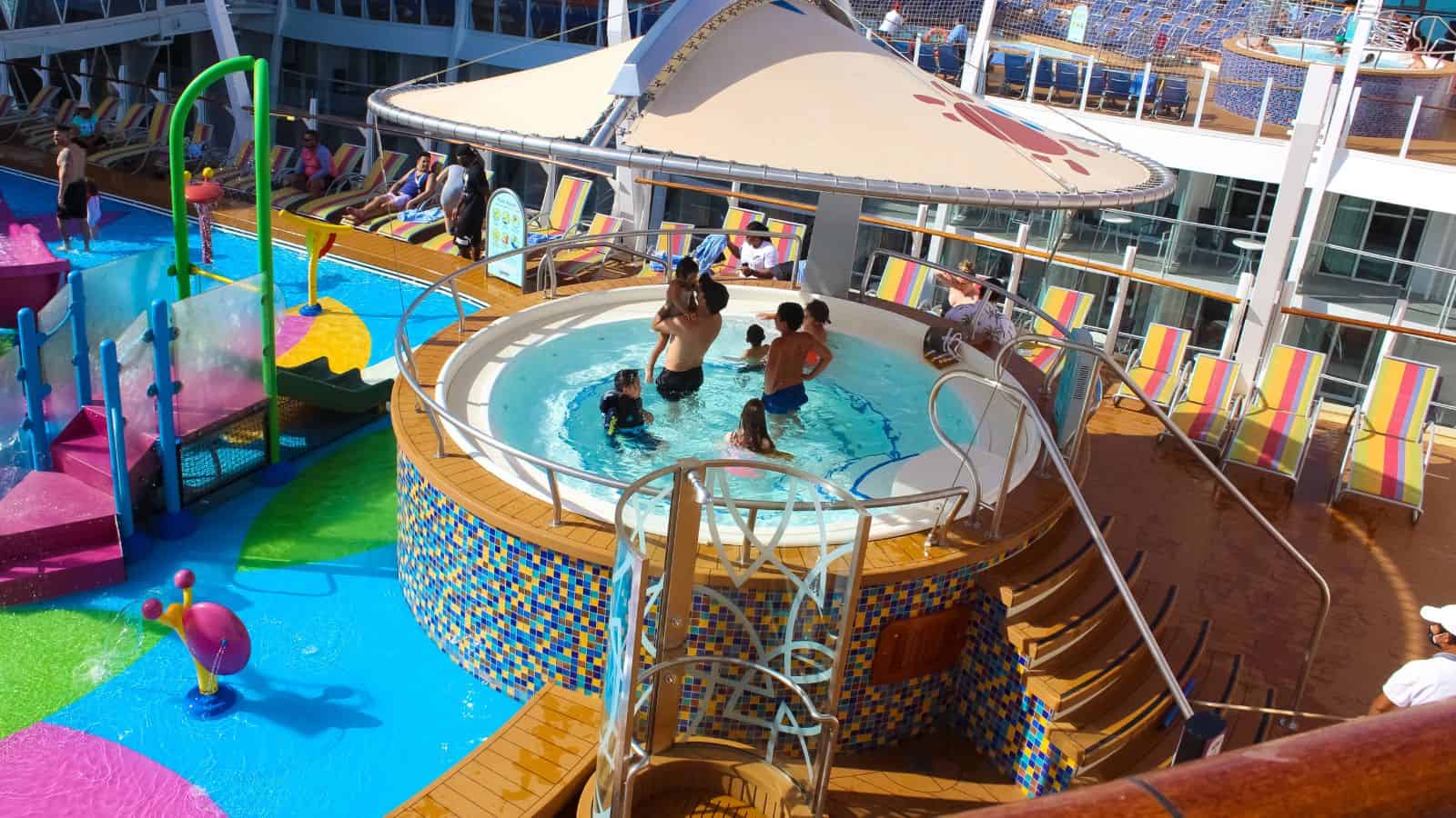 Several people are in a circular hot tub on a ship’s deck near a colorful splash pad and lounge chairs, with others sitting or walking nearby under a large canopy.