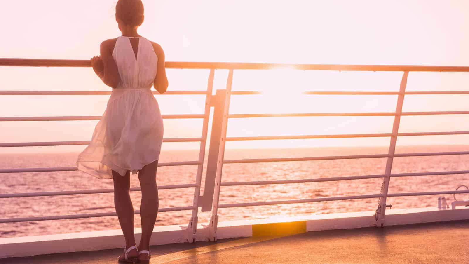 A person in a white dress stands on the deck of a ship, looking out at the ocean during sunset. The sunlight reflects on the water, and the scene has a warm, serene atmosphere.