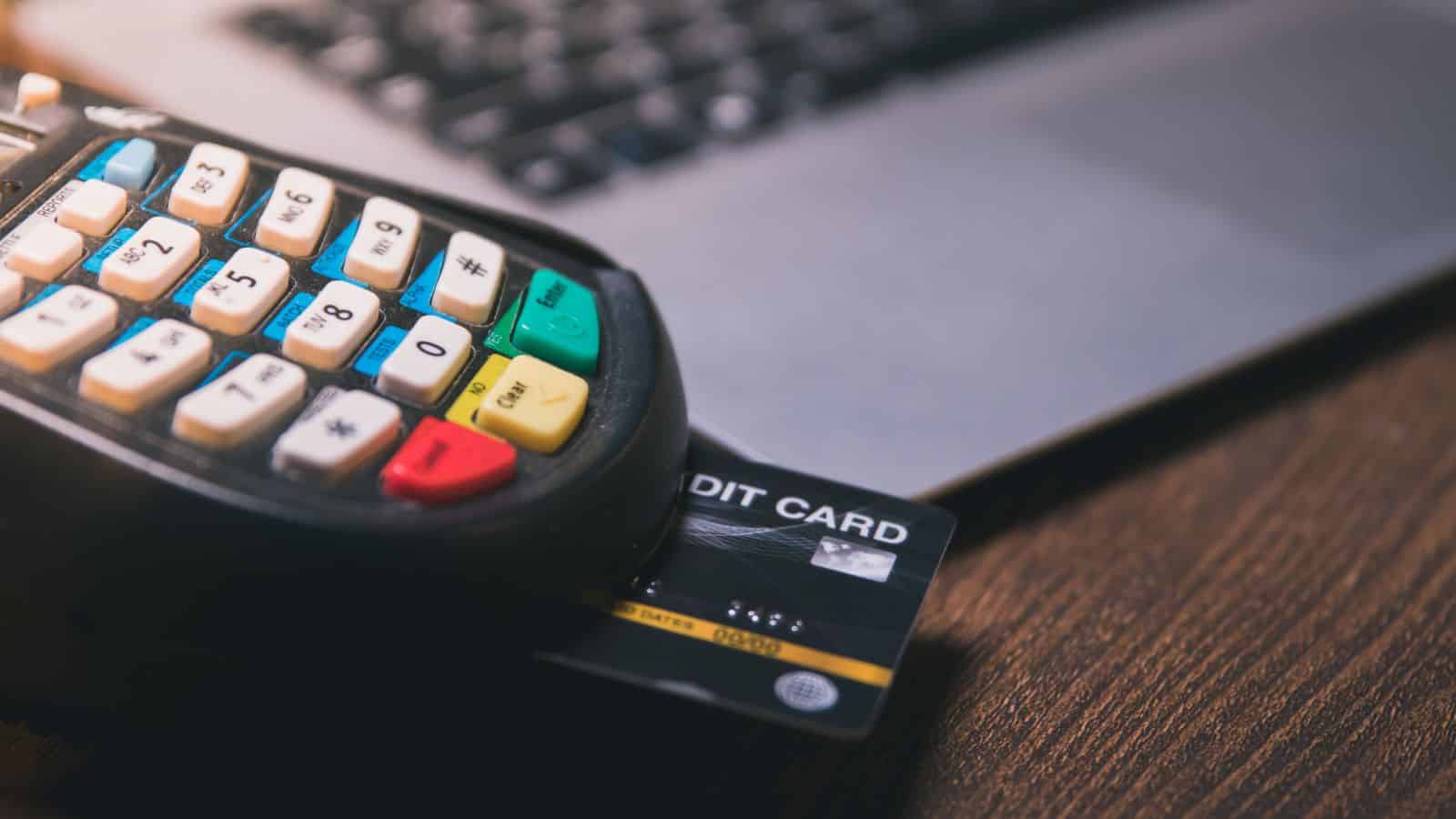 A close-up of a credit card inserted into a payment terminal, with a laptop in the background, all placed on a wooden surface.