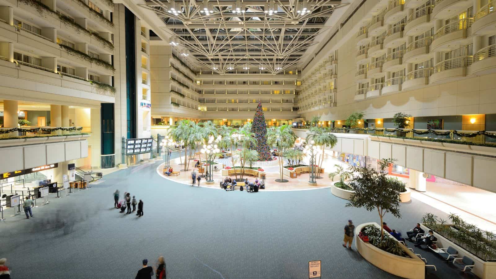 Spacious airport atrium with high ceilings, a large decorated Christmas tree in the center, palm trees, several people sitting or walking, and hotel balconies overlooking the area.