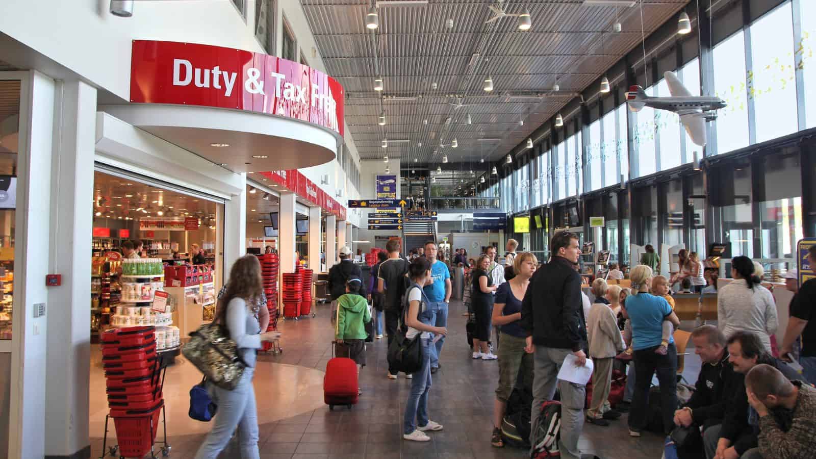 Travelers wait in a busy airport terminal near a Duty & Tax Free store. Some people stand with luggage and baskets, while others sit along the wall. Large windows line one side, letting in natural light.