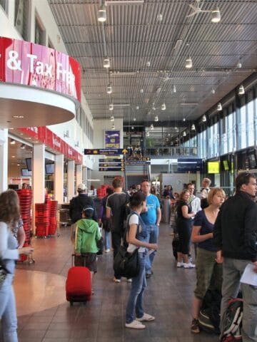Travelers wait in a busy airport terminal near a Duty & Tax Free store. Some people stand with luggage and baskets, while others sit along the wall. Large windows line one side, letting in natural light.