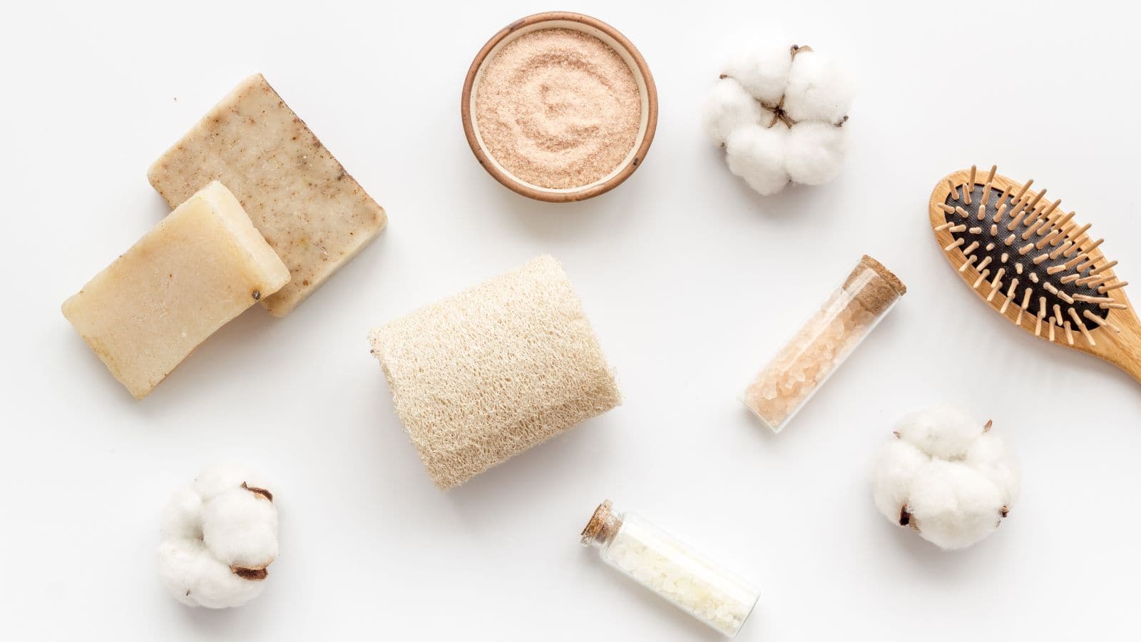 Various spa and self-care items, including bars of soap, a loofah, a wooden hairbrush, small jars of bath salts, cotton flowers, and a bowl of powder, are arranged on a white background.