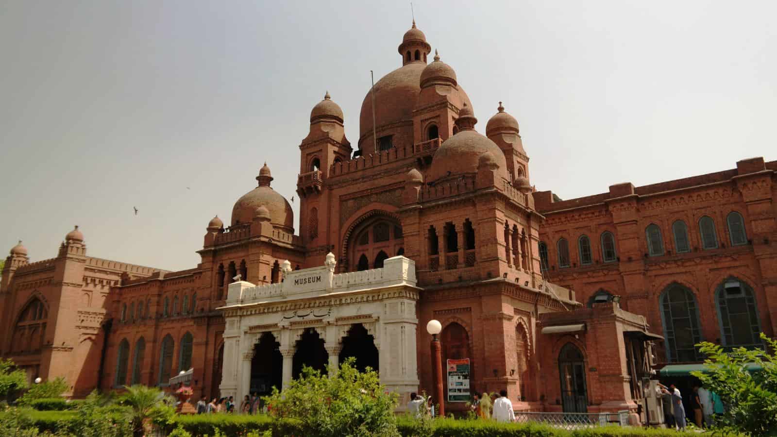A large, red-brick building with domes and arches, housing a museum. People are gathered near the entrance, and greenery surrounds the structure. The sky above is clear.