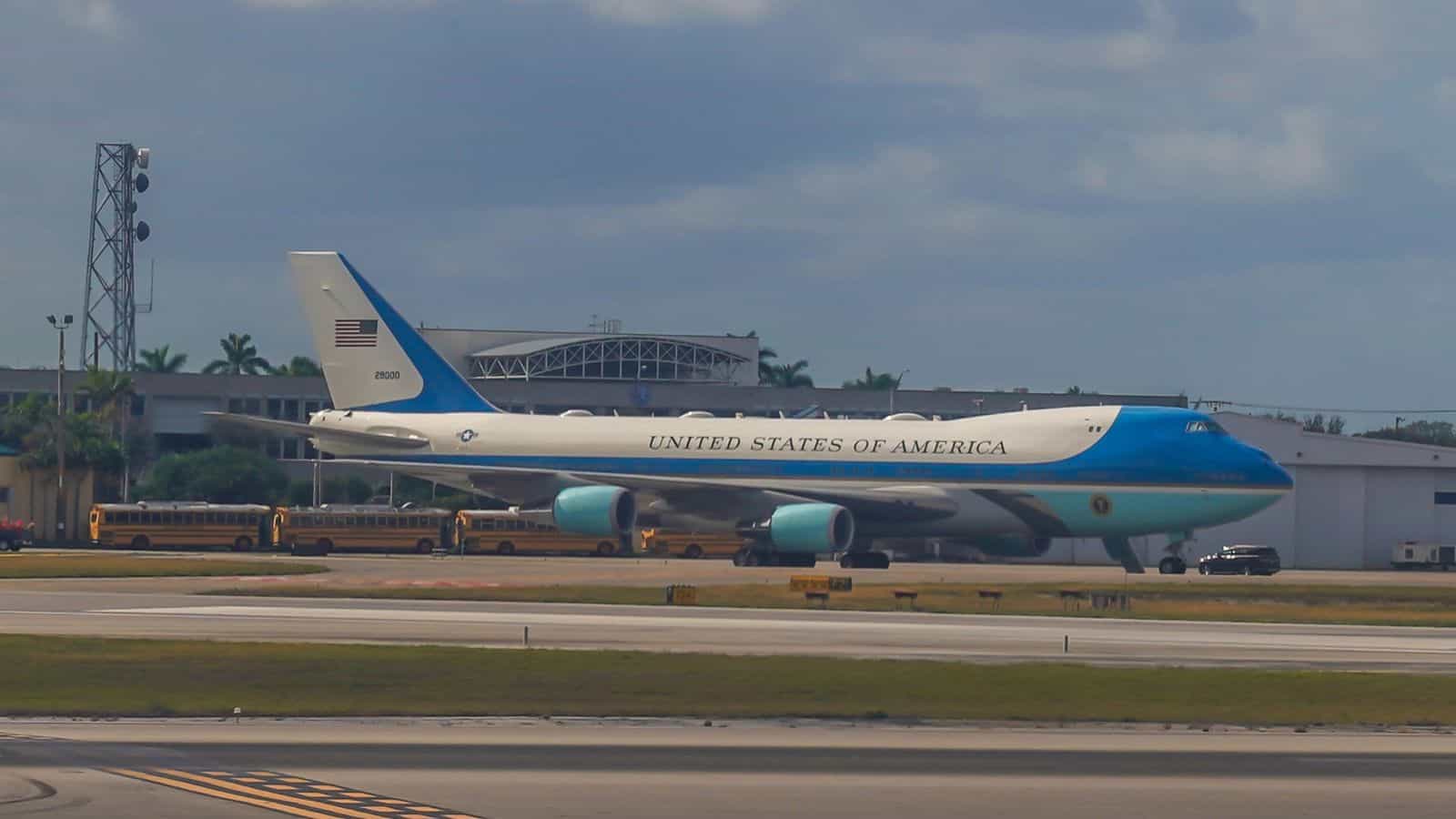 A large blue and white aircraft with "United States of America" painted on the side is parked on an airport tarmac, with several yellow school buses and a hangar visible in the background.