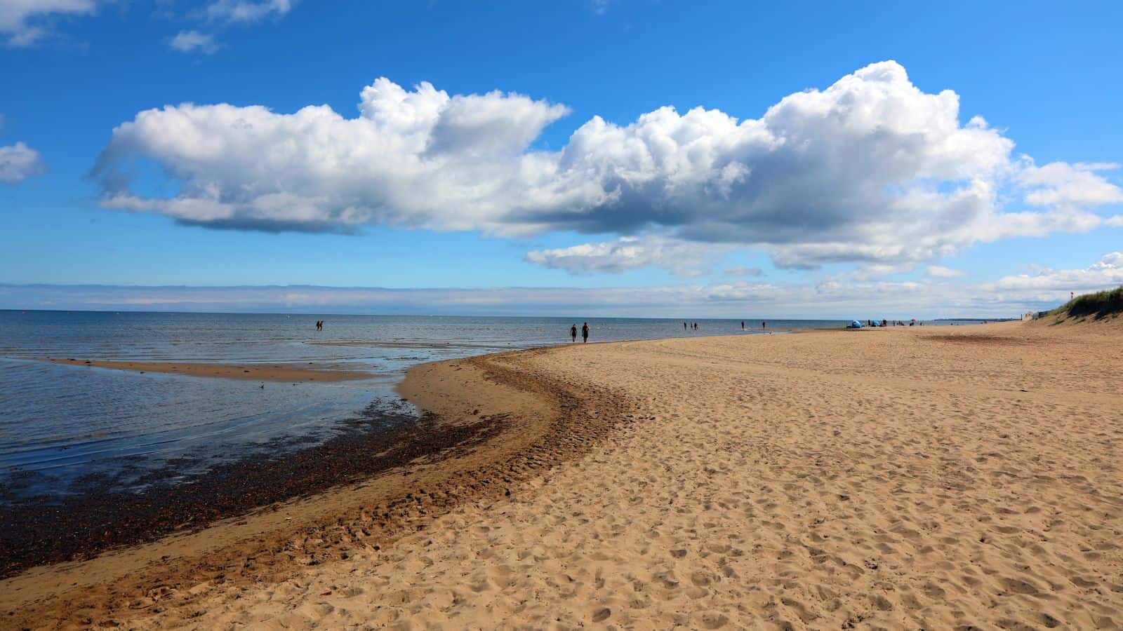 A sandy beach curves along the shoreline under a blue sky with large white clouds. A few people walk in the distance near the water’s edge. The sea is calm, and some grassy dunes are visible on the right.