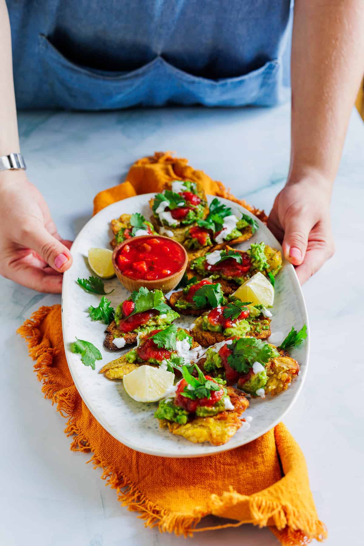 Person holding a plate of fritters topped with salsa, herbs, lime wedges, and a bowl of red dipping sauce—showcasing traditional vegetarian Colombian dishes.
