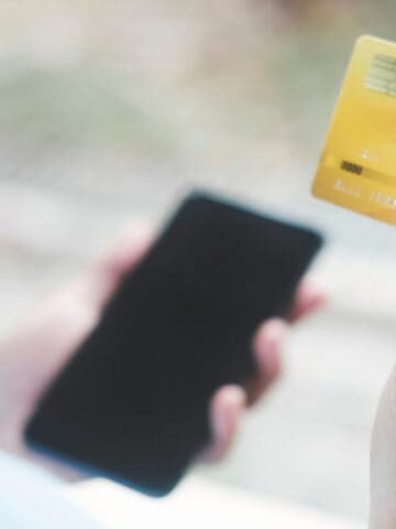 A person holds a yellow credit card in one hand and a smartphone in the other hand, possibly preparing to make an online payment or purchase. The background is blurred.