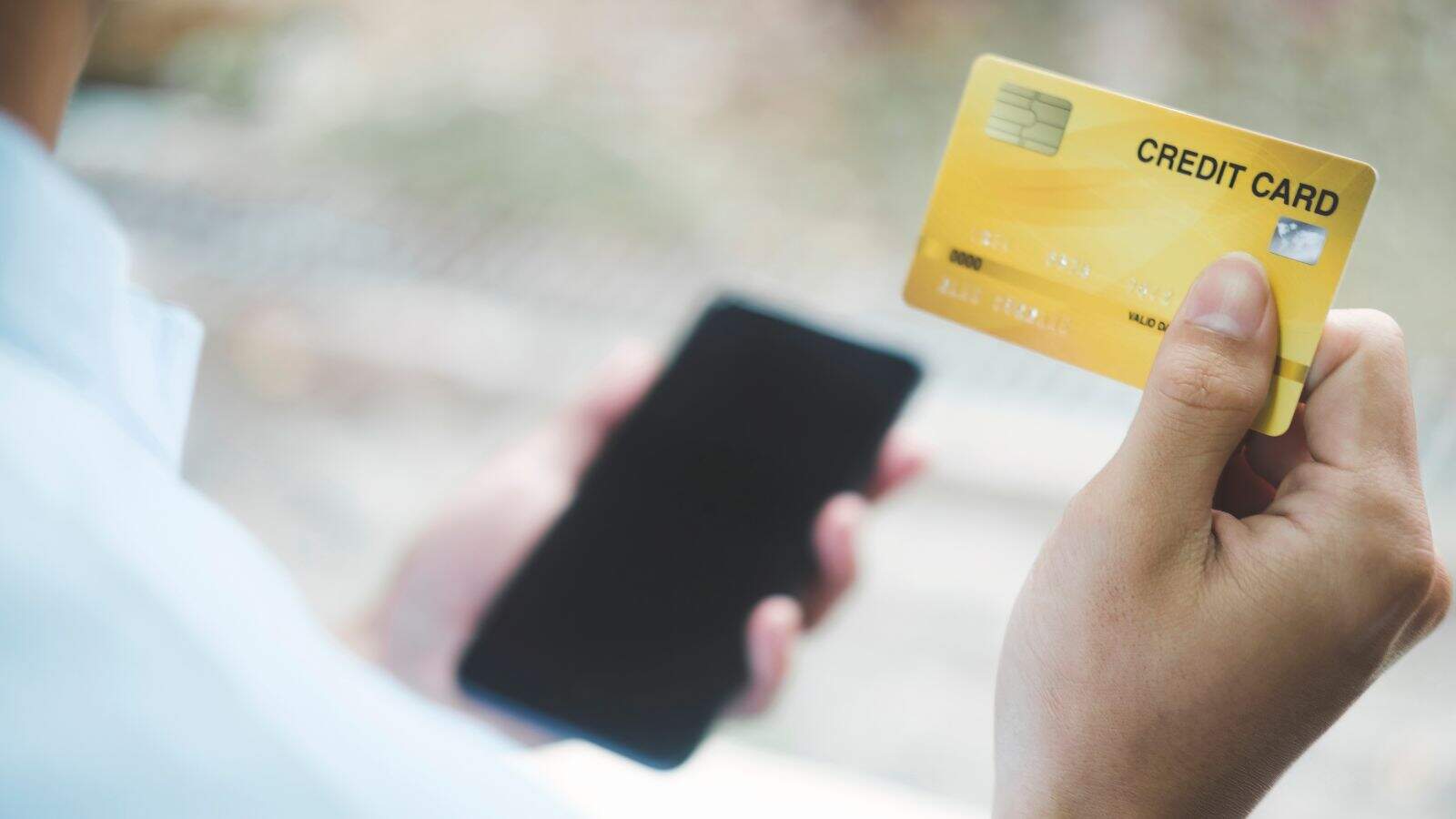 A person holds a yellow credit card in one hand and a smartphone in the other hand, possibly preparing to make an online payment or purchase. The background is blurred.