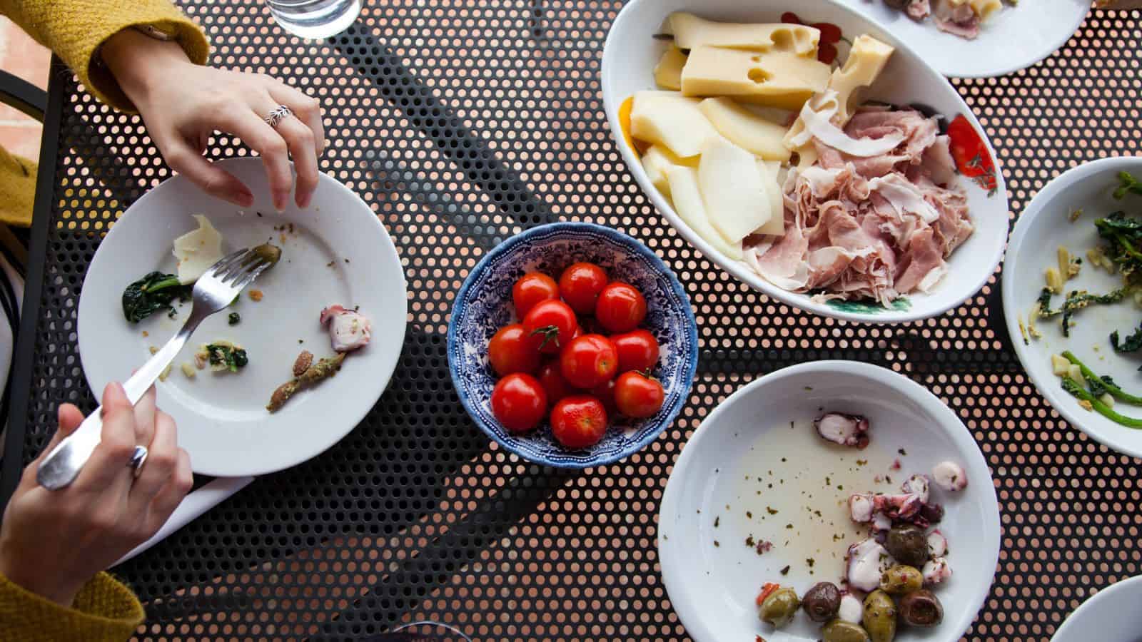 A table with plates of assorted food, including cherry tomatoes, cheese, and cold cuts. Two Travelers in Europe are visible&mdash;one using a fork and knife, the other reaching for food&mdash;sharing a meal amidst travel challenges.