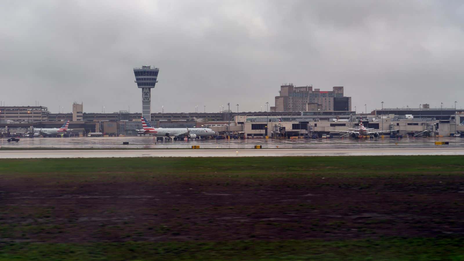 A view of an airport terminal with several airplanes parked at gates, a control tower, and a large building in the background under a cloudy, overcast sky. The foreground shows wet grass and pavement.