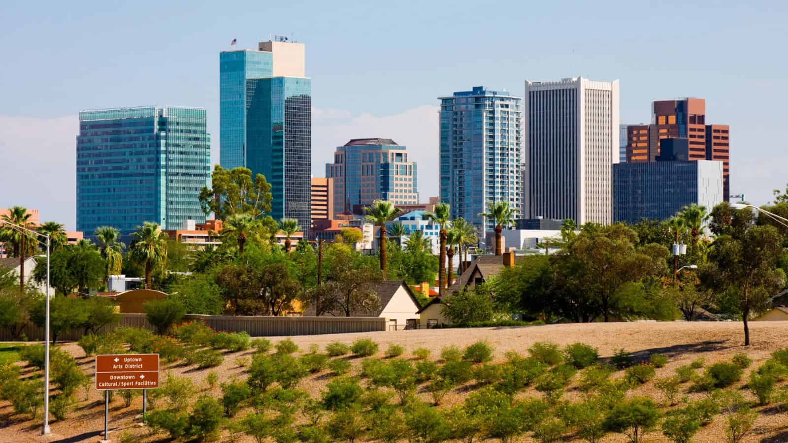 A city skyline with modern high-rise buildings and office towers behind a foreground of trees, shrubs, and a brown directional sign. The sky is clear and blue.