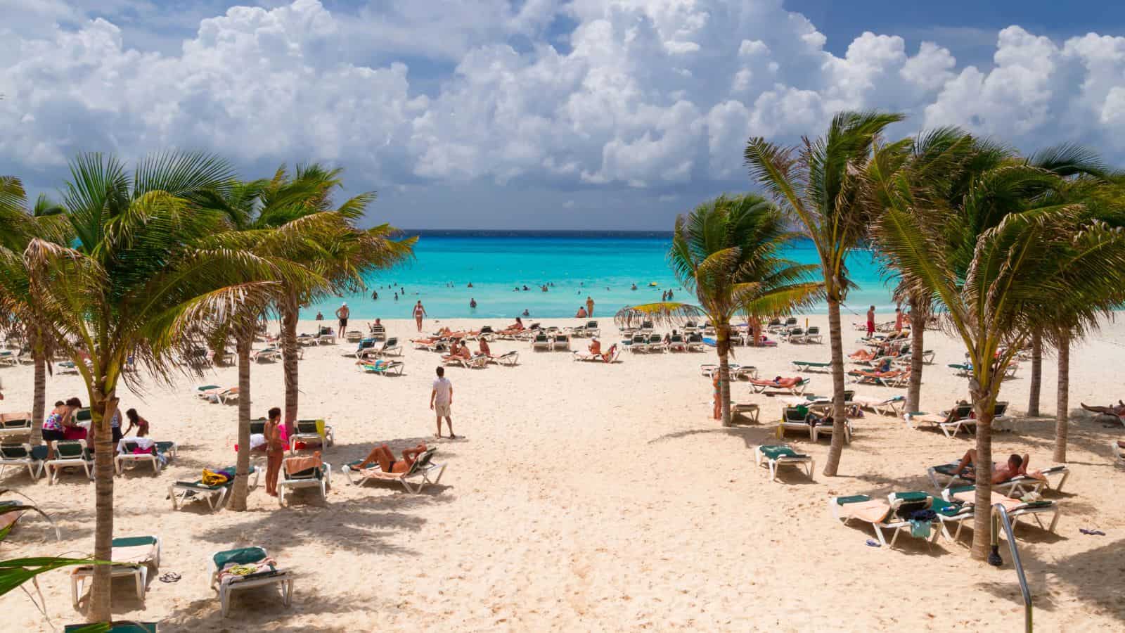 People relax on lounge chairs under palm trees on a sandy beach with turquoise water and scattered swimmers. The sky is partly cloudy, and the scene appears calm and tropical.