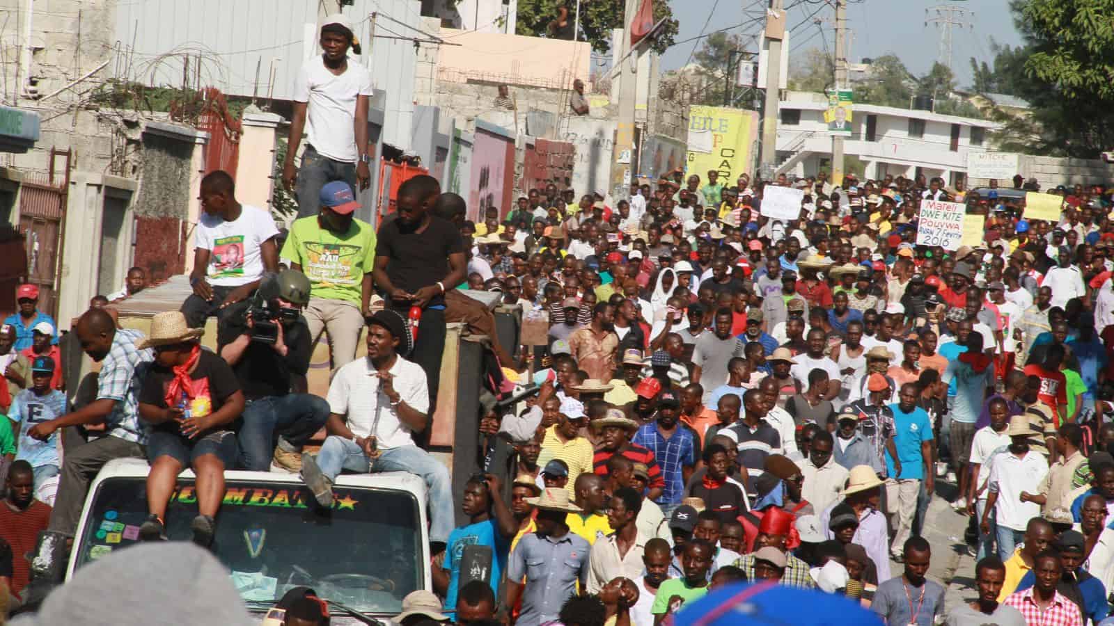 A large crowd of people marches down a city street during a protest; some ride on top of vehicles, while others carry signs. Buildings line the street, and the scene appears lively and densely packed.
