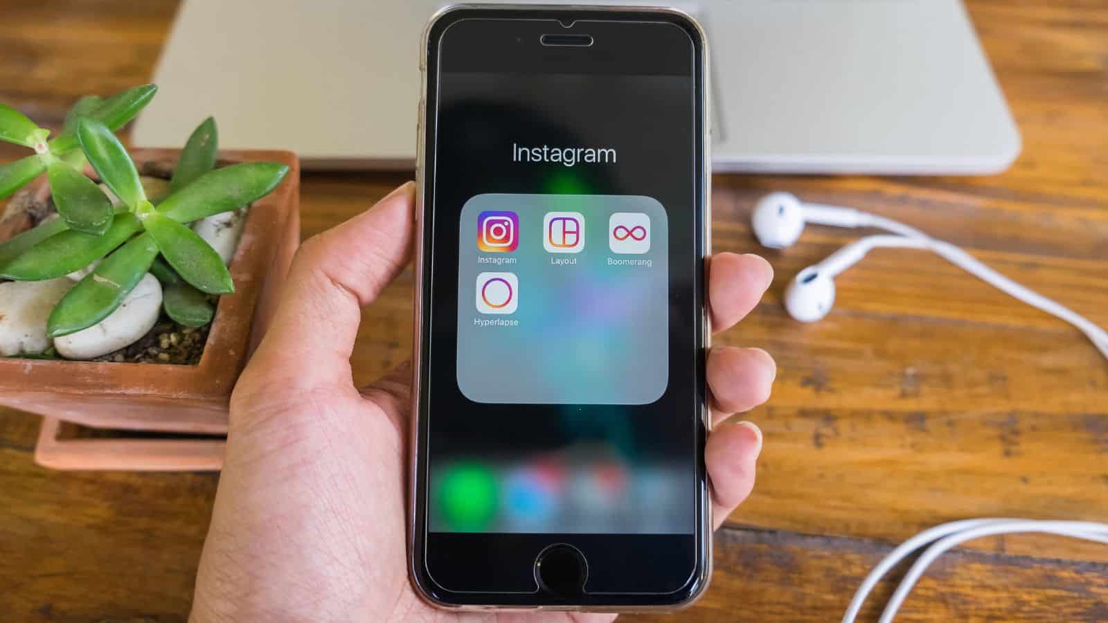 A hand holds a smartphone displaying a folder with Instagram-related apps. A potted succulent, laptop, and white earphones are on a wooden table in the background.