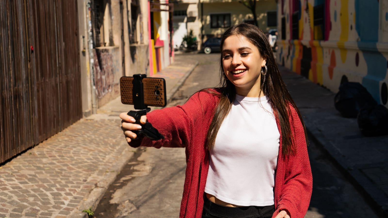 A woman with long brown hair wearing a white shirt and red cardigan smiles while holding a smartphone on a small tripod, taking a selfie in a sunlit, colorful alleyway.