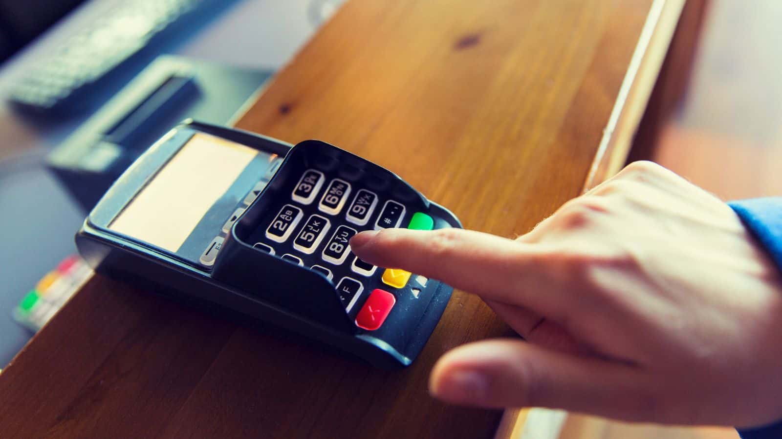 A person’s hand entering numbers on a payment terminal keypad, likely entering a PIN, with the device placed on a wooden counter.