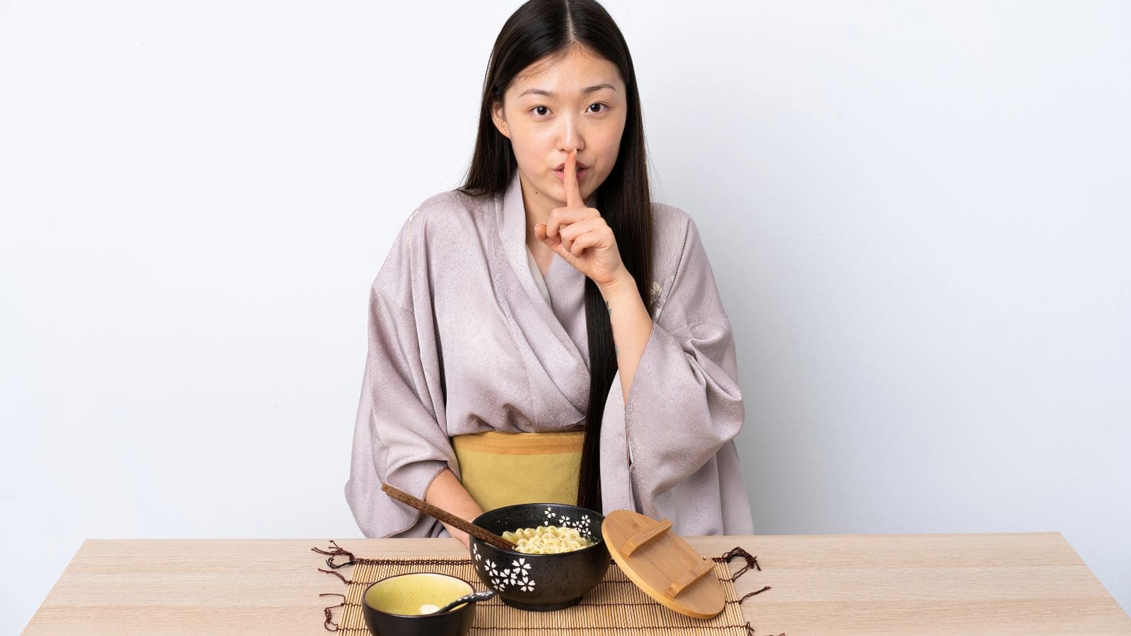 A woman in a kimono sits at a table with a bowl of rice and a wooden serving spoon, holding one finger to her lips in a shushing gesture.