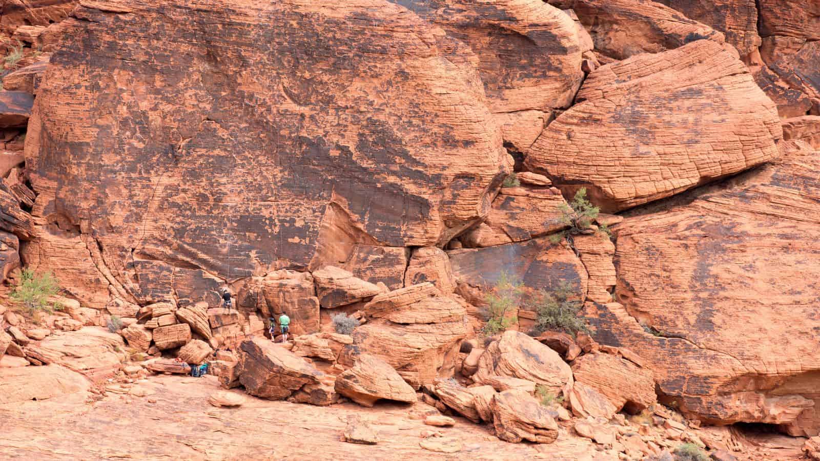 Three people stand at the base of a large, reddish-brown rock formation with visible layers and cracks, surrounded by scattered rocks and sparse vegetation.