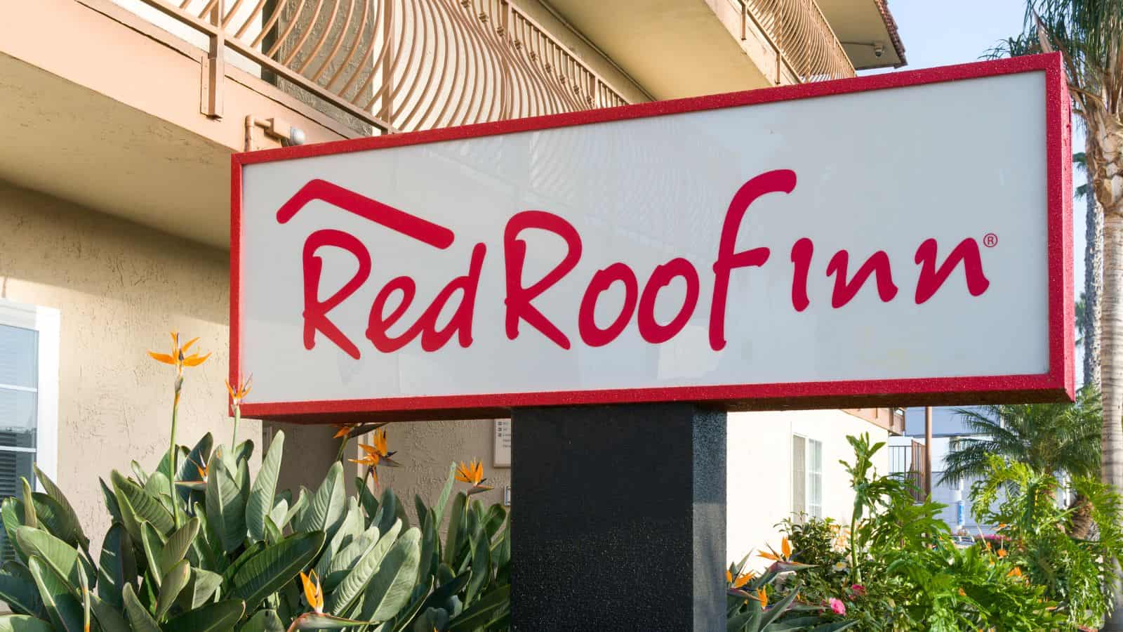 A Red Roof Inn sign in front of a beige building with balconies, surrounded by green plants and orange flowers—one of the US hotel chains disappointing guests recently.