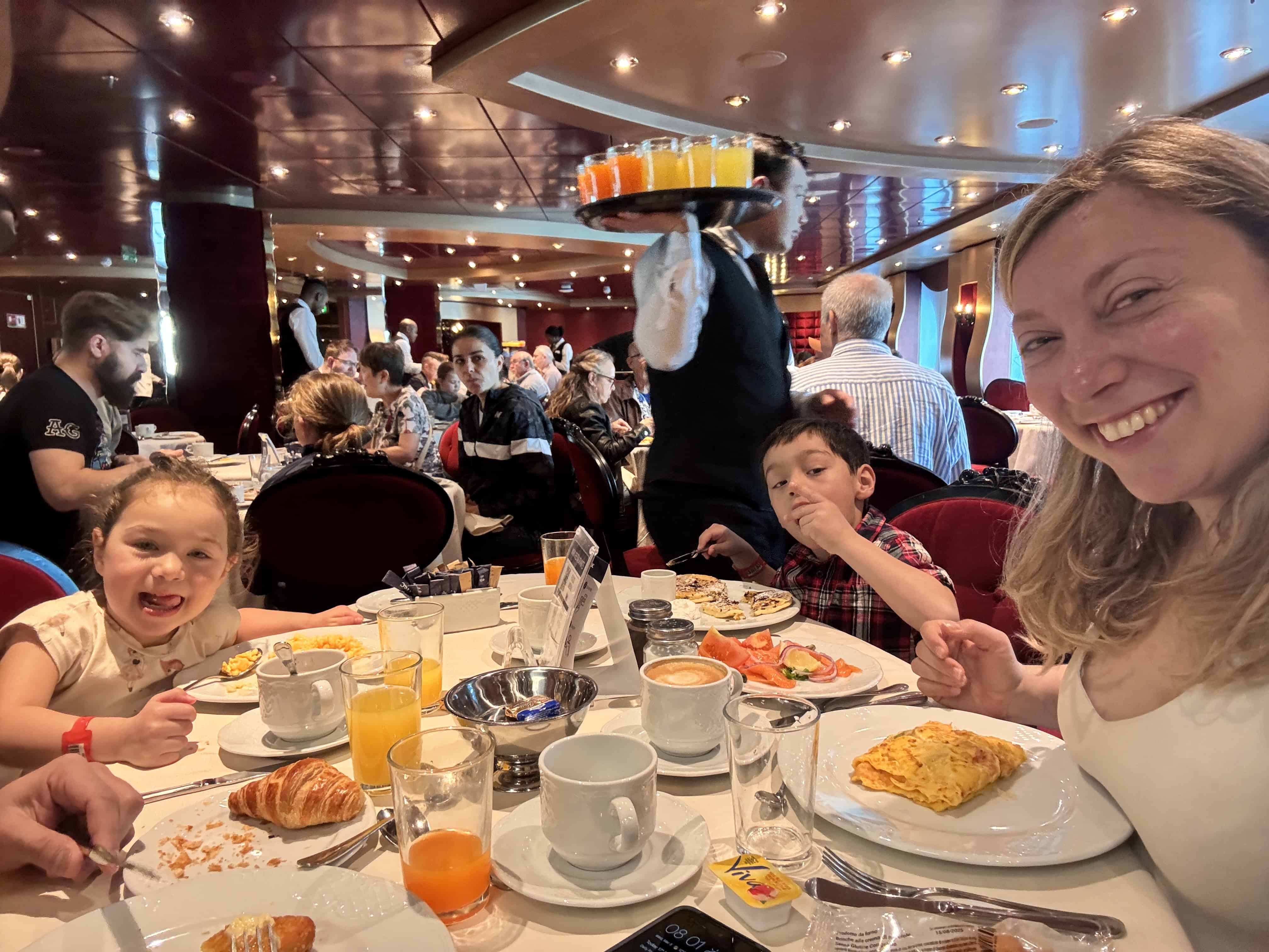 A woman and two children smile while having breakfast in a busy restaurant aboard MSC Fantasia Cruise, enjoying a memorable family cruise experience as a server carries drinks.